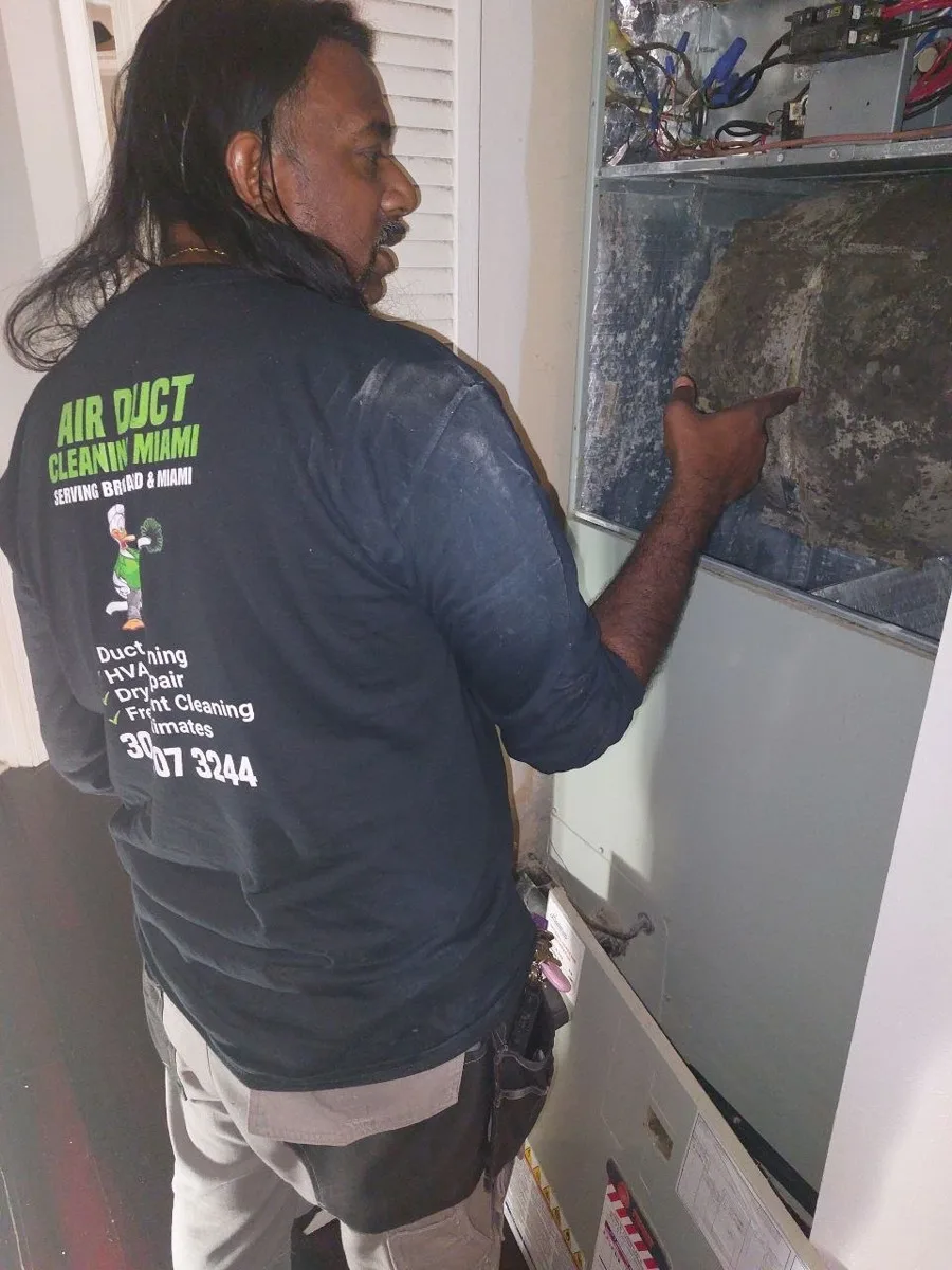 Air Duct Cleaning Miami technician in a branded uniform pointing at a blower fan and evaporator coil completely covered in mold inside a Florida air handler, before AC maintenance
