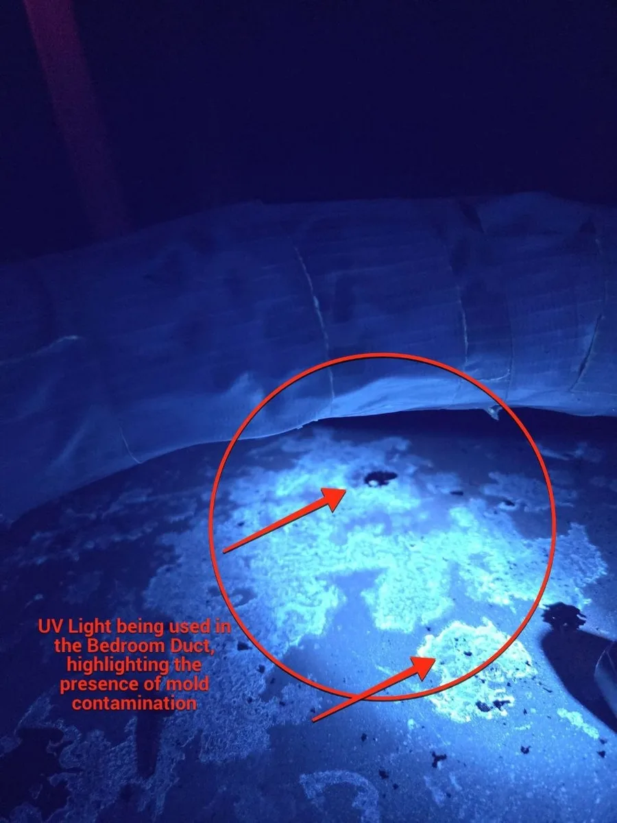 Bedroom flex duct and plenum under a UV lamp during a mold inspection, mold contamination fluorescing pale blue-white across a wide patch