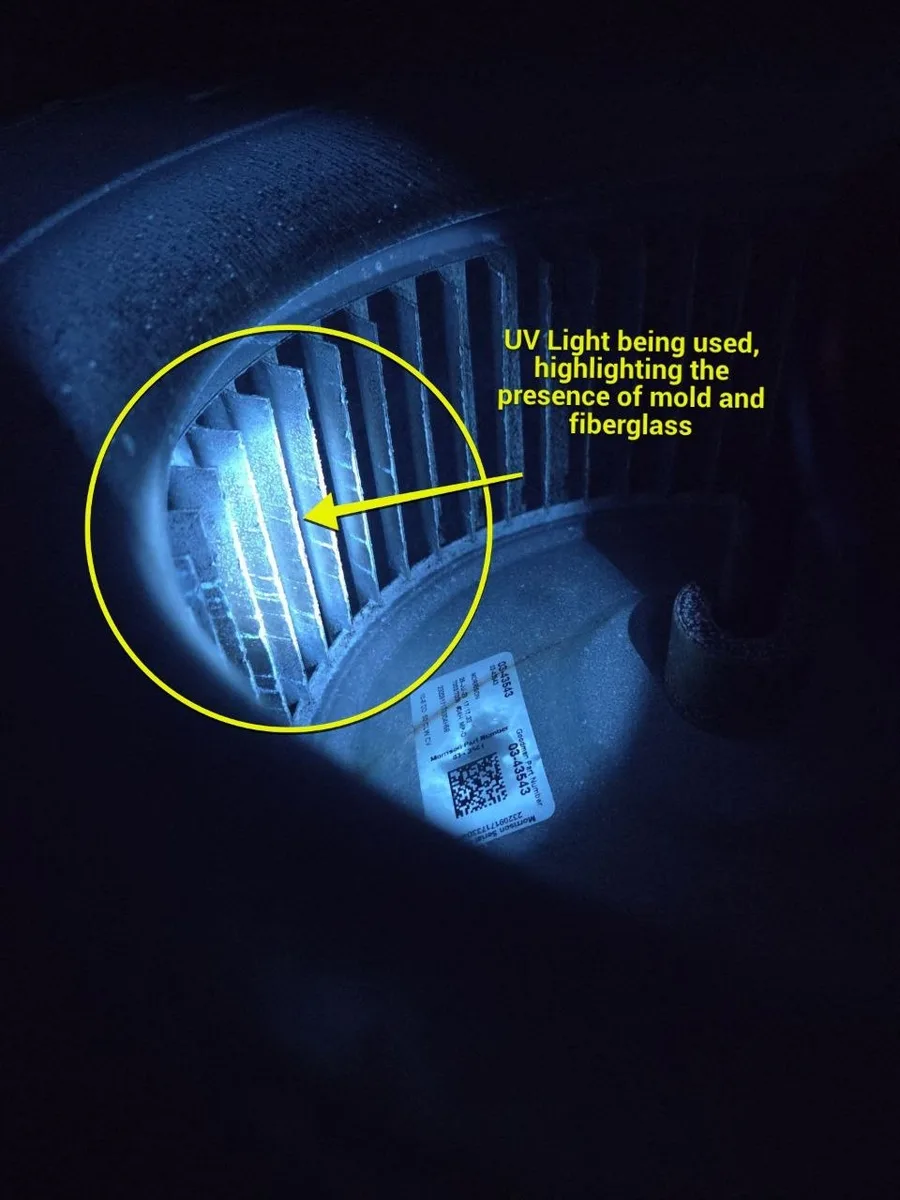 Air handler blower wheel under a UV lamp, fins lit up showing mold contamination and loose fiberglass, during a Miami-Dade duct mold inspection